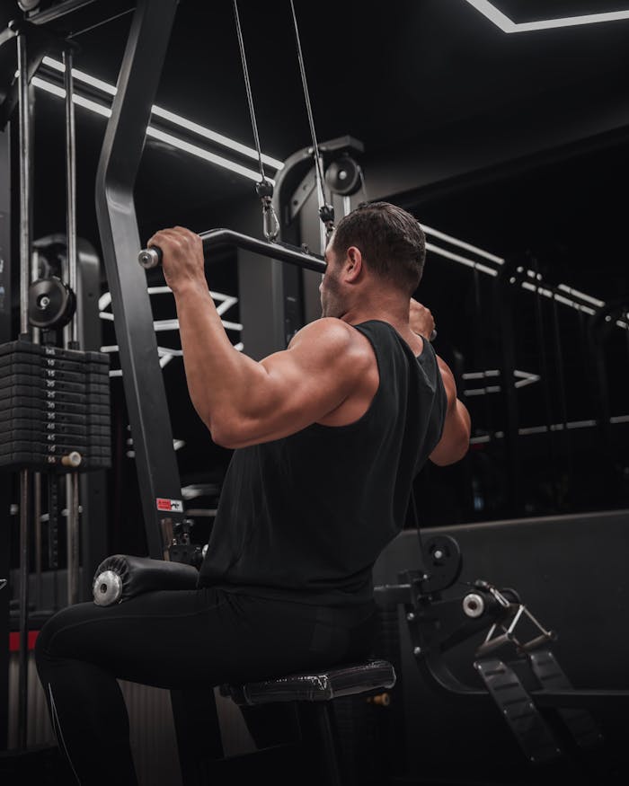 gallery-4 A muscular man performs a lat pulldown exercise on gym equipment, showcasing strength.